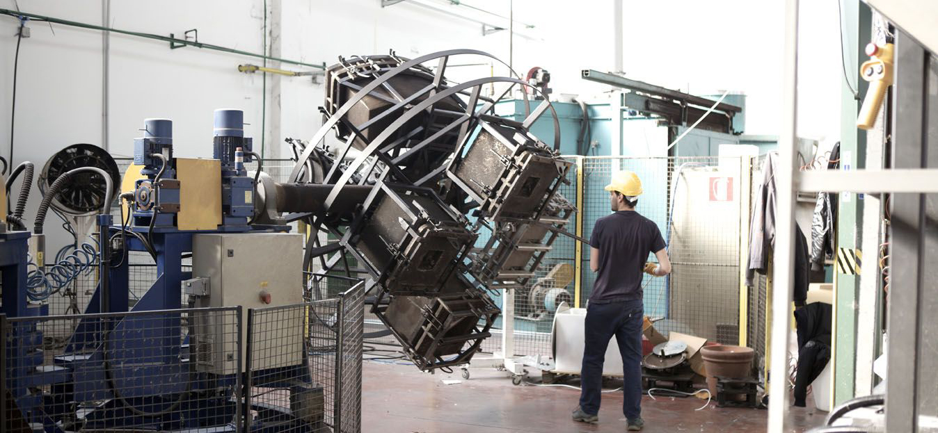 A worker in a manufacturing facility overseeing a large rotomolding machine. The machine is rotating a mold frame on two perpendicular axes, creating uniform plastic cases. The worker is wearing a yellow hard hat and is standing near machinery with safety barriers in place.